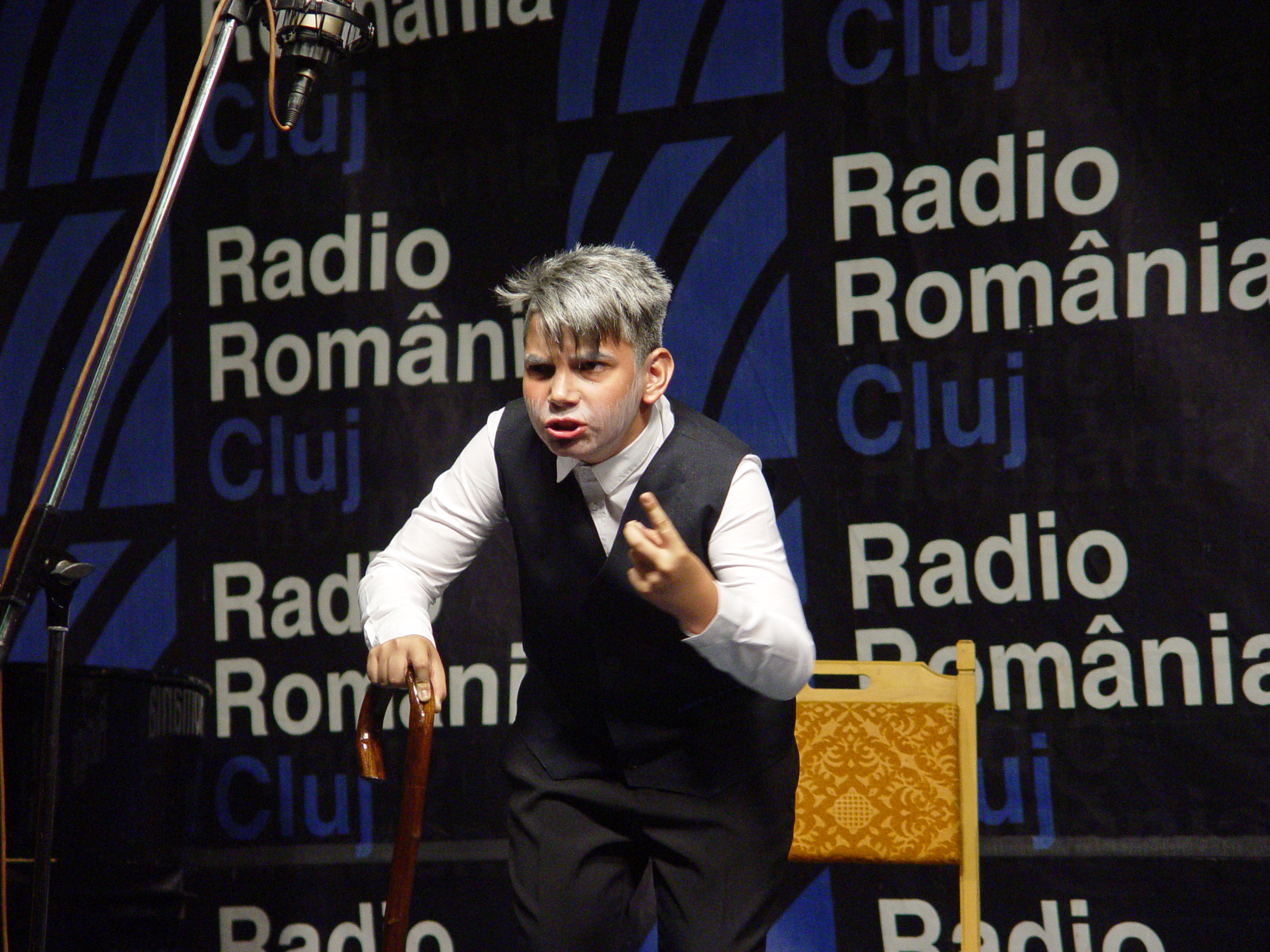 man wearing a black jacket, white shirt, blue jeans, and brown boots, playing a white electric guitar while sitting on an amp